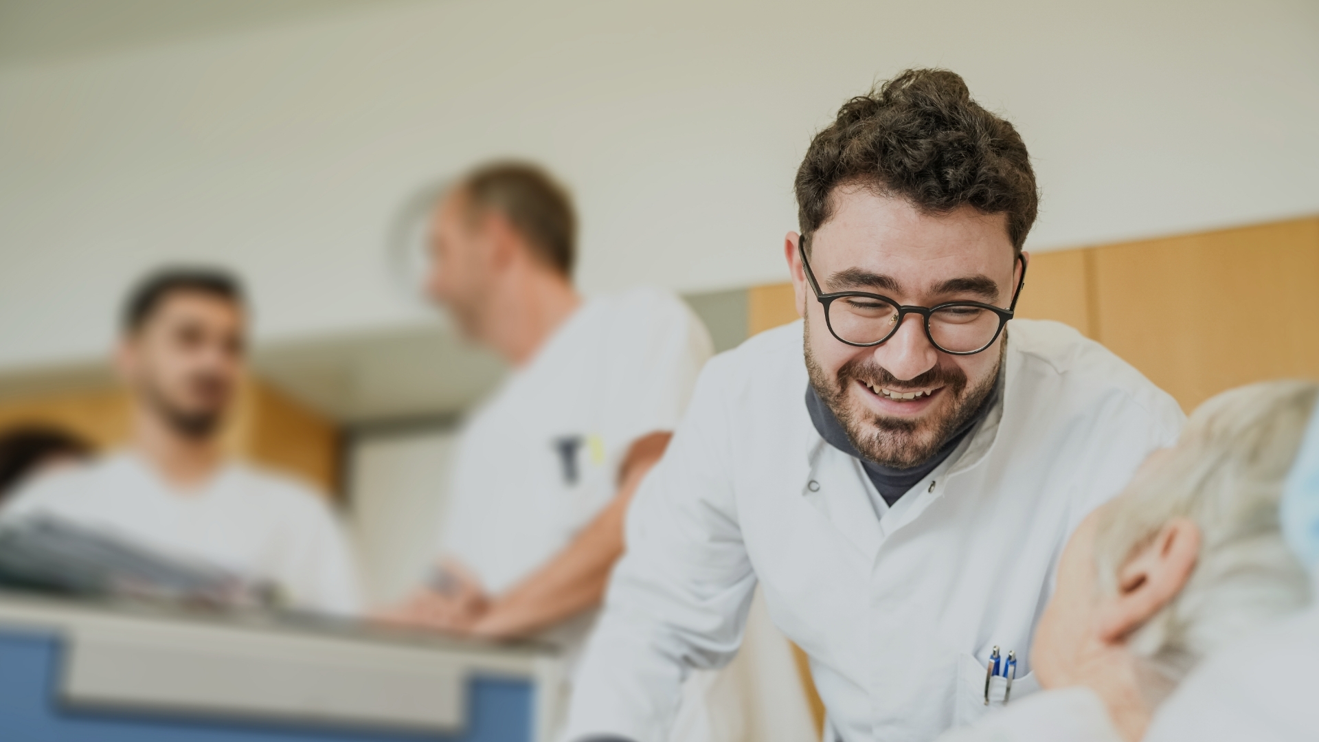 Arzt mit dunkler Brille und Vollbart beugt sich über älteren, liegenden Patienten, links im Bild zwei Kollegen verschwommen